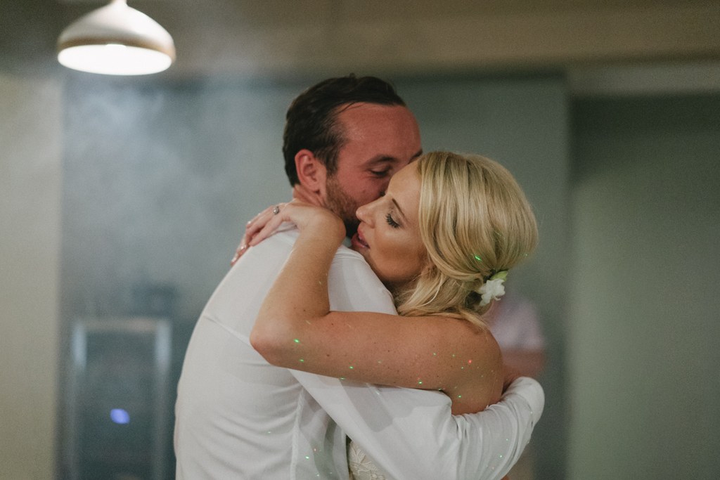 Groom in white shirt kisses bride — she wears a white gown and sparkle-decorated arm, warm light