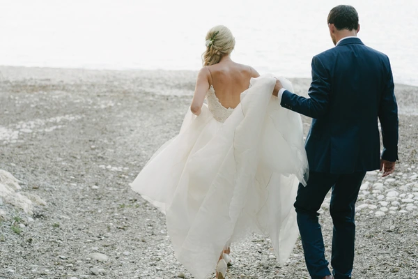 Bride and groom walk on a pebble beach, the white wedding dress catches the breeze