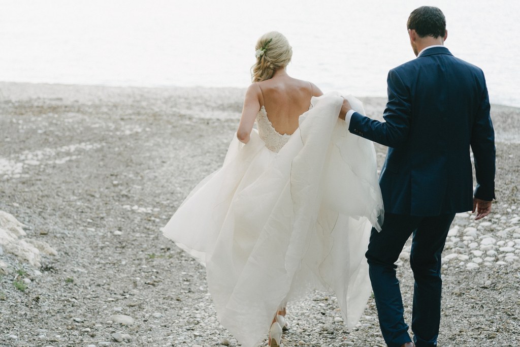 Bride and groom walk on a pebble beach, the white wedding dress catches the breeze