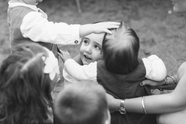 Black and white close-up of a toddler boy looking up with wide curious eyes, surrounded by children's hands reaching around him at the wedding. Candid documentary portrait.