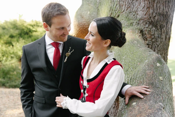 Bride in her Norwegian bunad smiling at the groom while leaning against a large boulder together in the Royal Botanic Garden Edinburgh. Relaxed couple portrait.