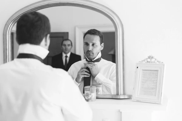 Black and white mirror reflection of the groom adjusting his tie while a groomsman watches from the background. Candid wedding morning detail.