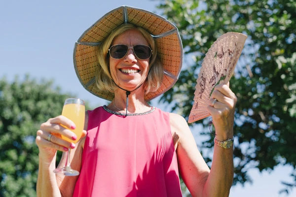 Smiling woman in pink top, wide hat and sunglasses holds champagne and a fan — green leaves above