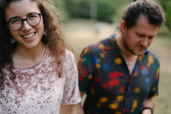 Young woman with glasses smiling at camera while man looks away in patterned shirt at wedding.