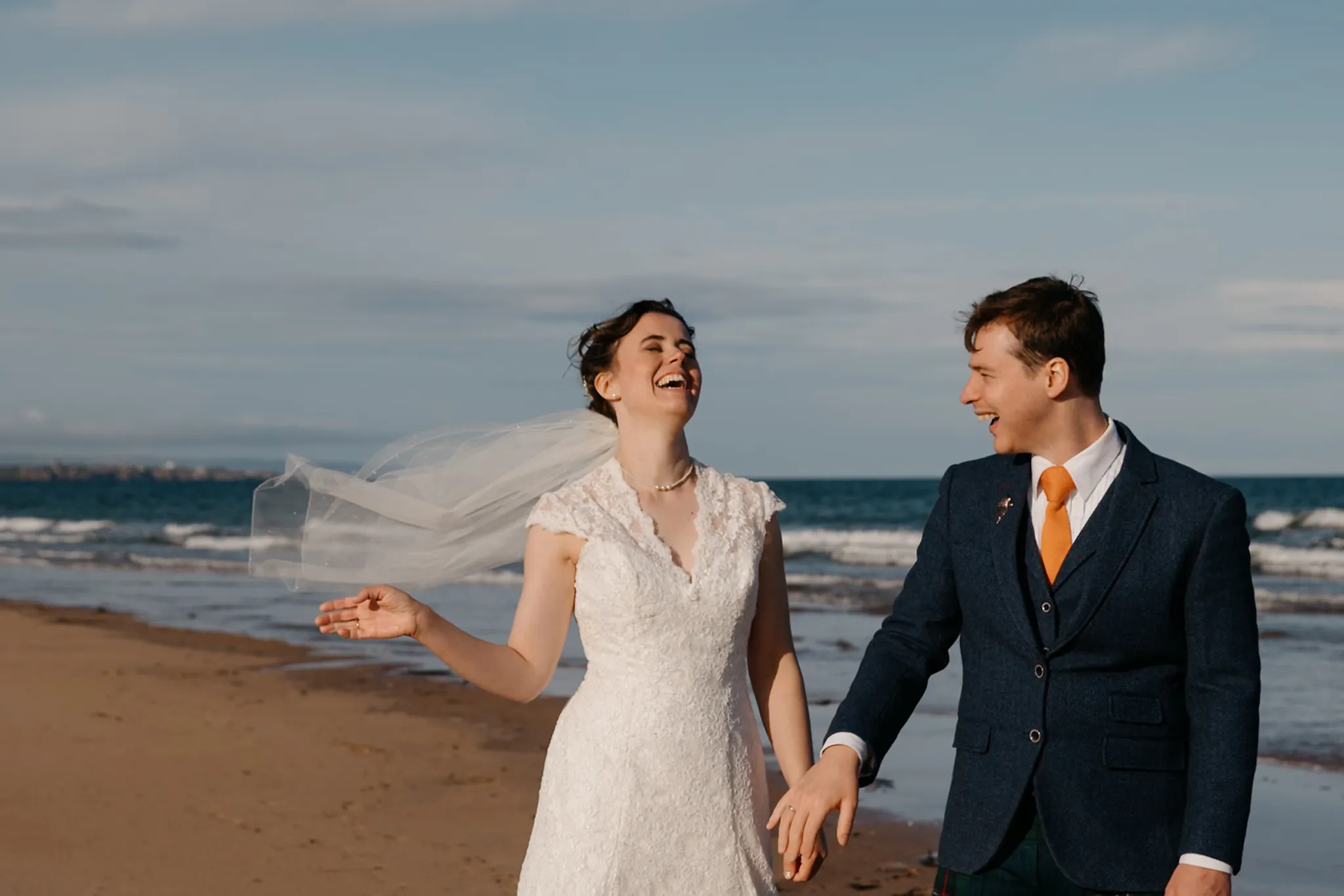 Laughing newlyweds walk hand-in-hand on a windy beach, bride's veil flying, groom in navy suit with orange tie.