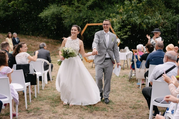 Bride and groom walking hand in hand past seated wedding guests under hexagon wooden arch.