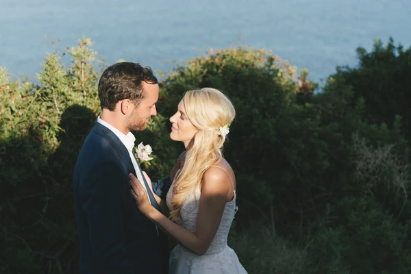 Bride and groom in intimate gaze — blue sea and green vegetation behind, warm afternoon light