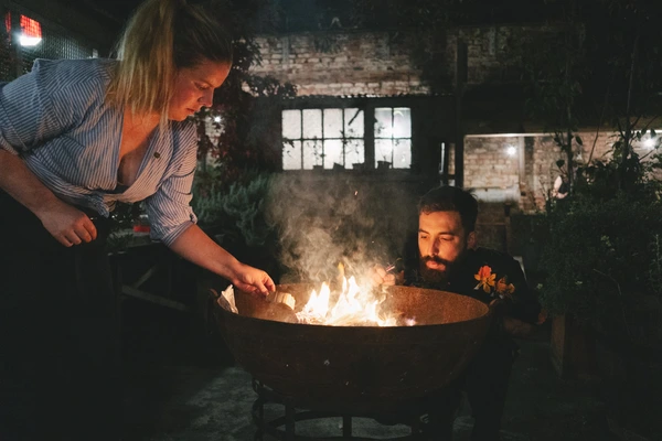 Bride and groom light a fire in a round fire pit at night — intimate moment, brick walls behind