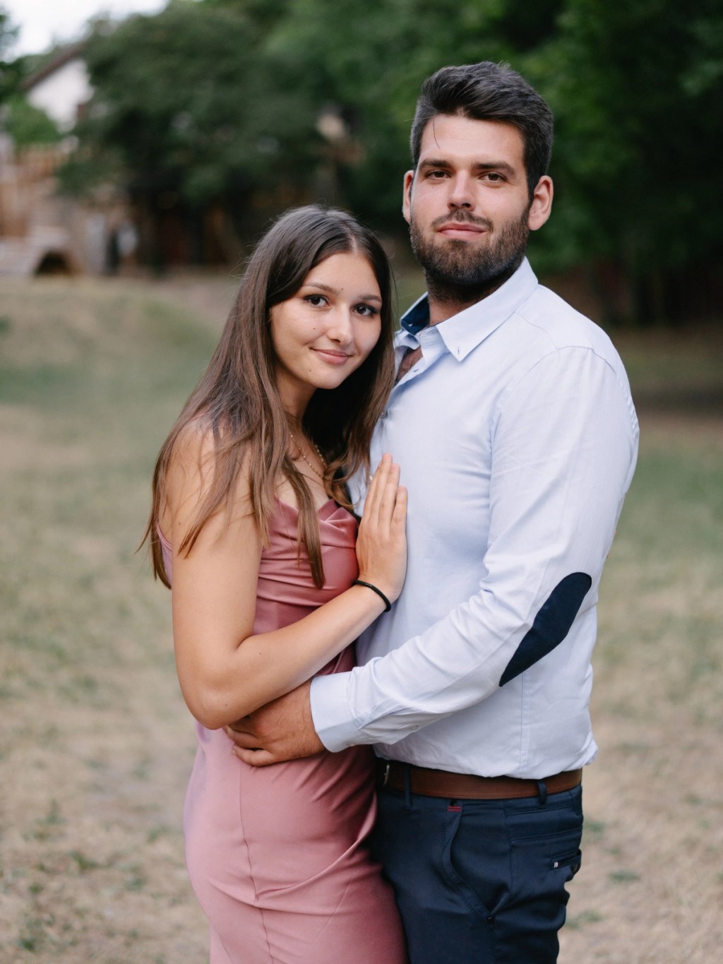 Couple posing together in pink and white outfit at outdoor wedding venue with green background.