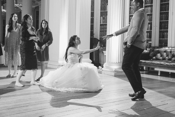 Bride kneels with raised hands toward the groom while family watches — elegant library-like room