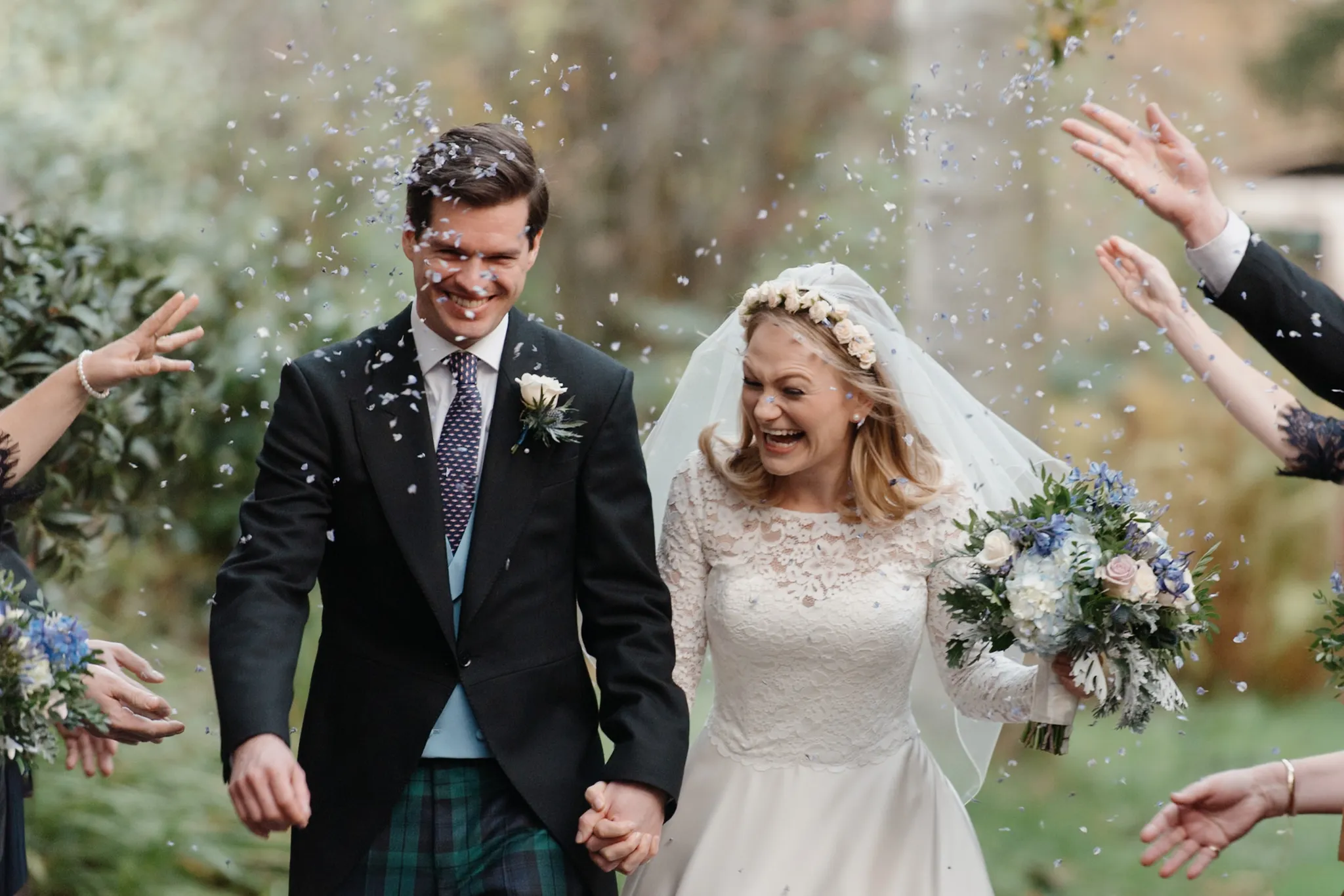 Newlyweds walking through confetti shower at outdoor wedding ceremony, bride in white lace dress and veil, groom in dark suit and tartan kilt.