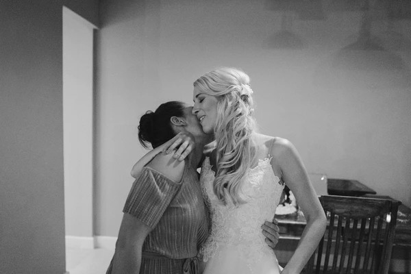 Bride with long wavy hair smiles as a dark-haired woman in pyjamas gives her a kiss on the cheek