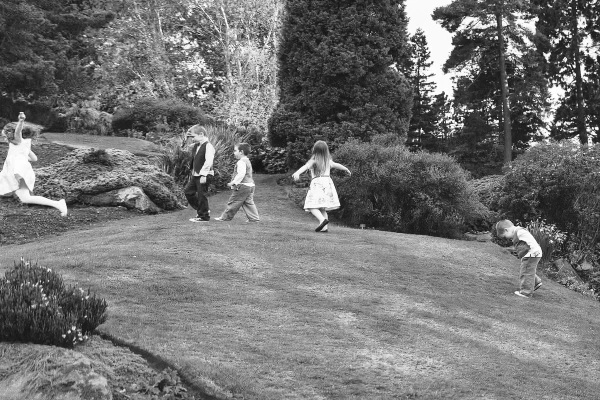 Black and white image of several children running and playing on a sloped grass lawn surrounded by shrubs and trees at the Royal Botanic Garden Edinburgh wedding. Candid documentary moment.