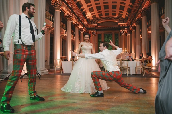 Smiling bride and groom in Scottish kilt dance in the baroque ballroom — orange-lit columns
