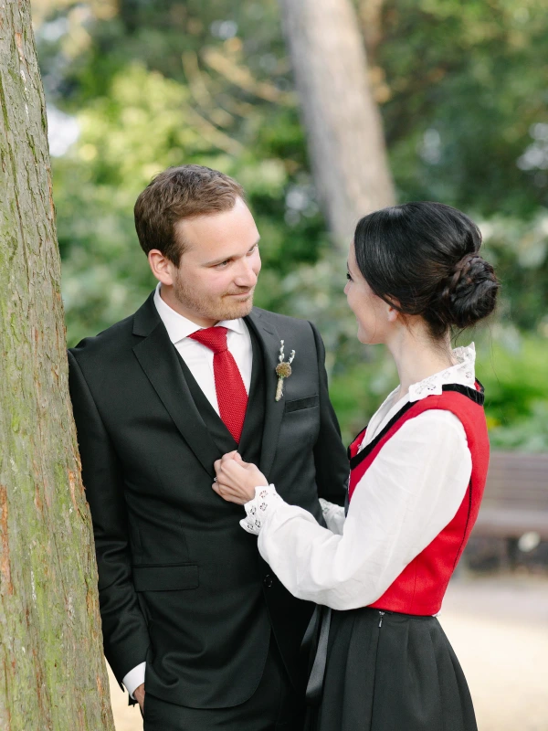 The bride in a Norwegian bunad looks up at the groom affectionately as she rests her hand on his chest beside a tree at the Royal Botanic Garden Edinburgh. Candid documentary wedding portrait.