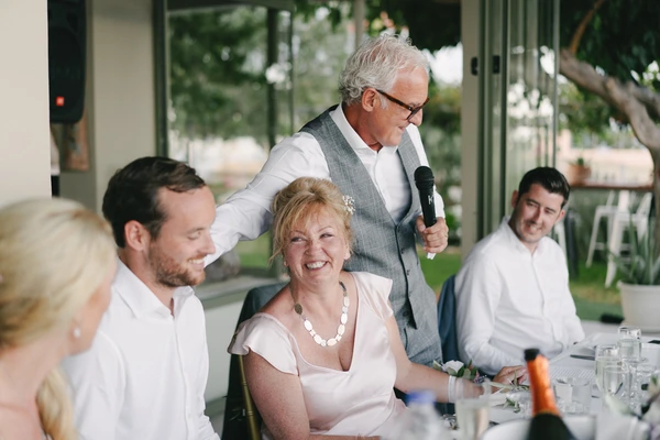 Older man in grey vest speaks at the wedding table, smiling mother in white with pearls beside him