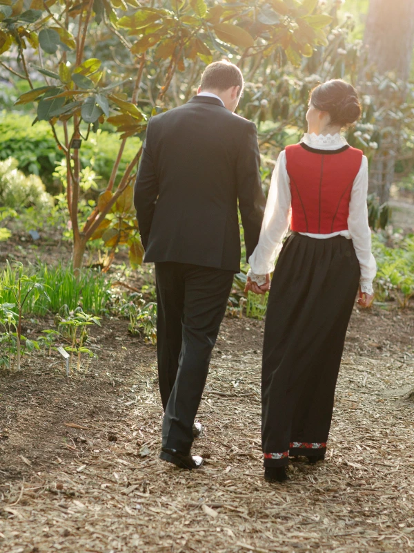 The bride in a Norwegian red and black bunad and the groom in a dark suit walk hand in hand through a sunlit garden, photographed from behind. Candid wedding portrait at the Royal Botanic Garden Edinburgh.