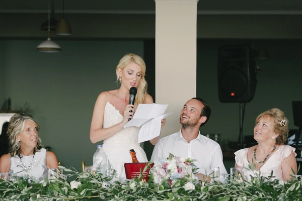 Bride speaks with a microphone at the dinner table in white satin, paper in hand, guests listening