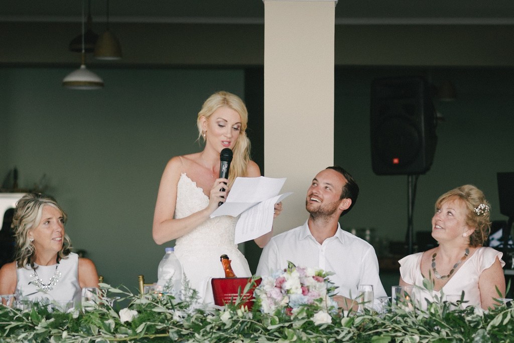 Bride speaks with a microphone at the dinner table in white satin, paper in hand, guests listening