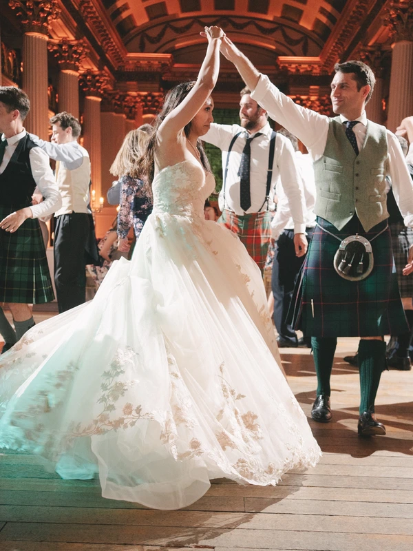 Bride in white lace dances with the groom in Scottish kilt — guests around, warm orange light