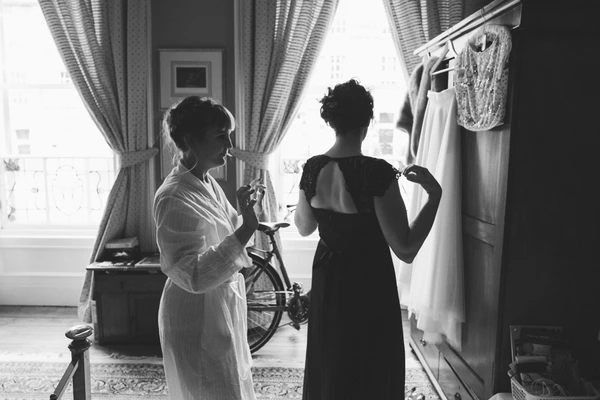 Two women in an elegant room during dress preparation — one in a white robe, one in black. Black-and-white intimate scene.