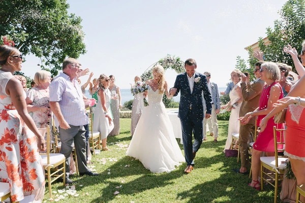 Newlyweds walk the aisle as guests shower them with flower petals — sea garden, sunny celebration