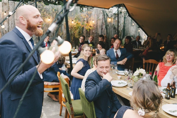 Groom gives a speech at the marquee reception, guests laughing and listening
