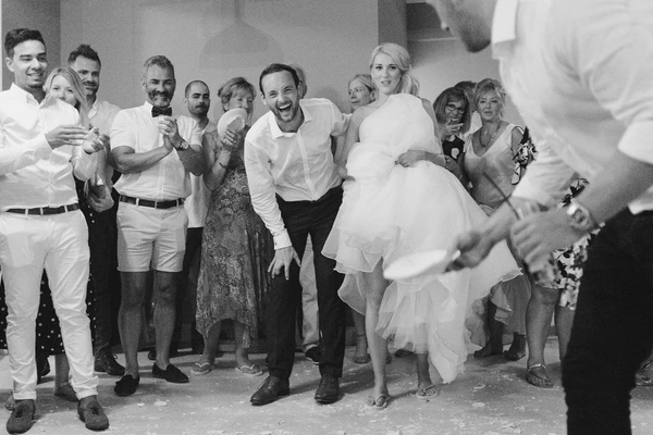 Groom laughs as bride's white dress flies up — family around them, joyful party. Black-and-white.