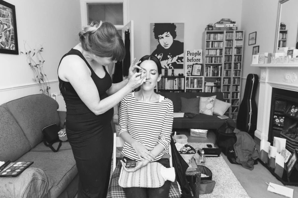 Black and white wide shot of the make-up artist working on the bride in a Victorian-style living room with bay windows and a Bob Dylan poster. Candid bridal preparations.