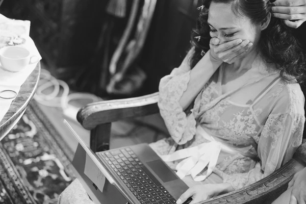 Bride in elegant lace covers her mouth in surprise while looking at the laptop — black-and-white