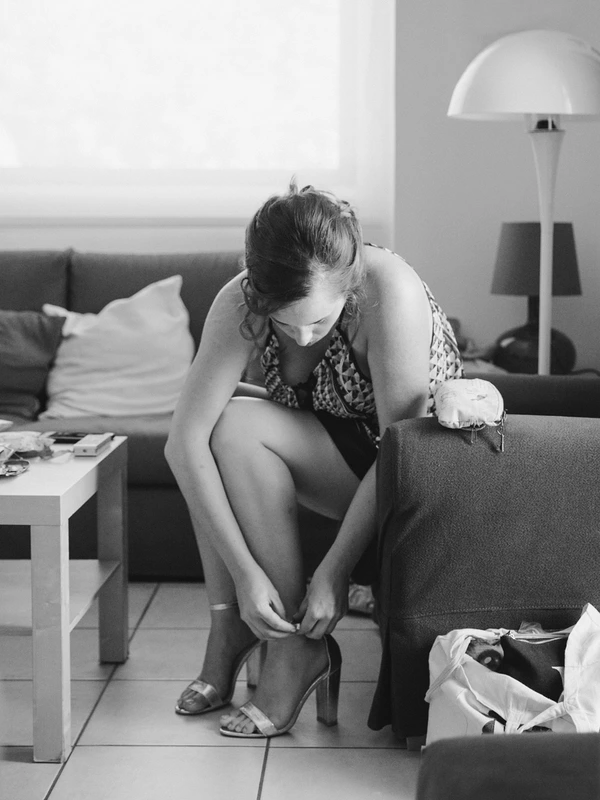 Bride pulls on her shoe on the sofa on the wedding morning — focused, elegant motion in modern room