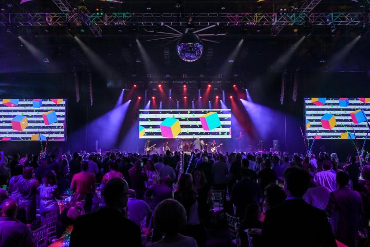 Transformational event lighting and video production are on display as an 80’s tribute band performs at a themed corporate event with large stage screens.