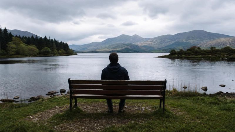 A person sitting alone on a bench looking out at a calm Irish lake, representing why loneliness sticks