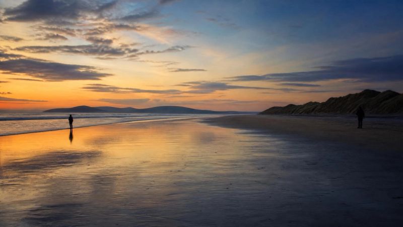 Two silhouettes on a beach at dusk, one closer to the water than the other