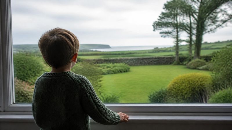 A child seen from behind standing at a window looking out at a green garden