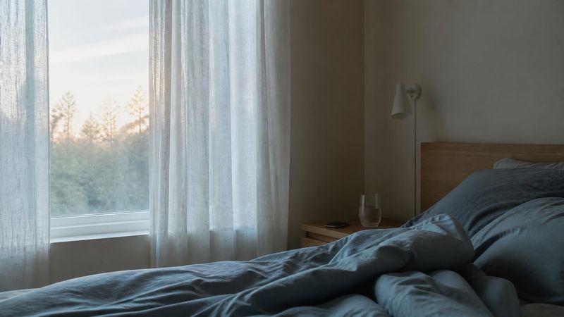 A bedroom at dawn with an unmade bed and pale morning light through curtains