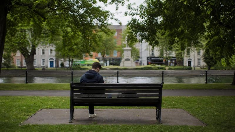 A person sitting alone on a park bench in Dublin, looking down at their phone, illustrating how adult ADHD symptoms are often hidden or misattributed.