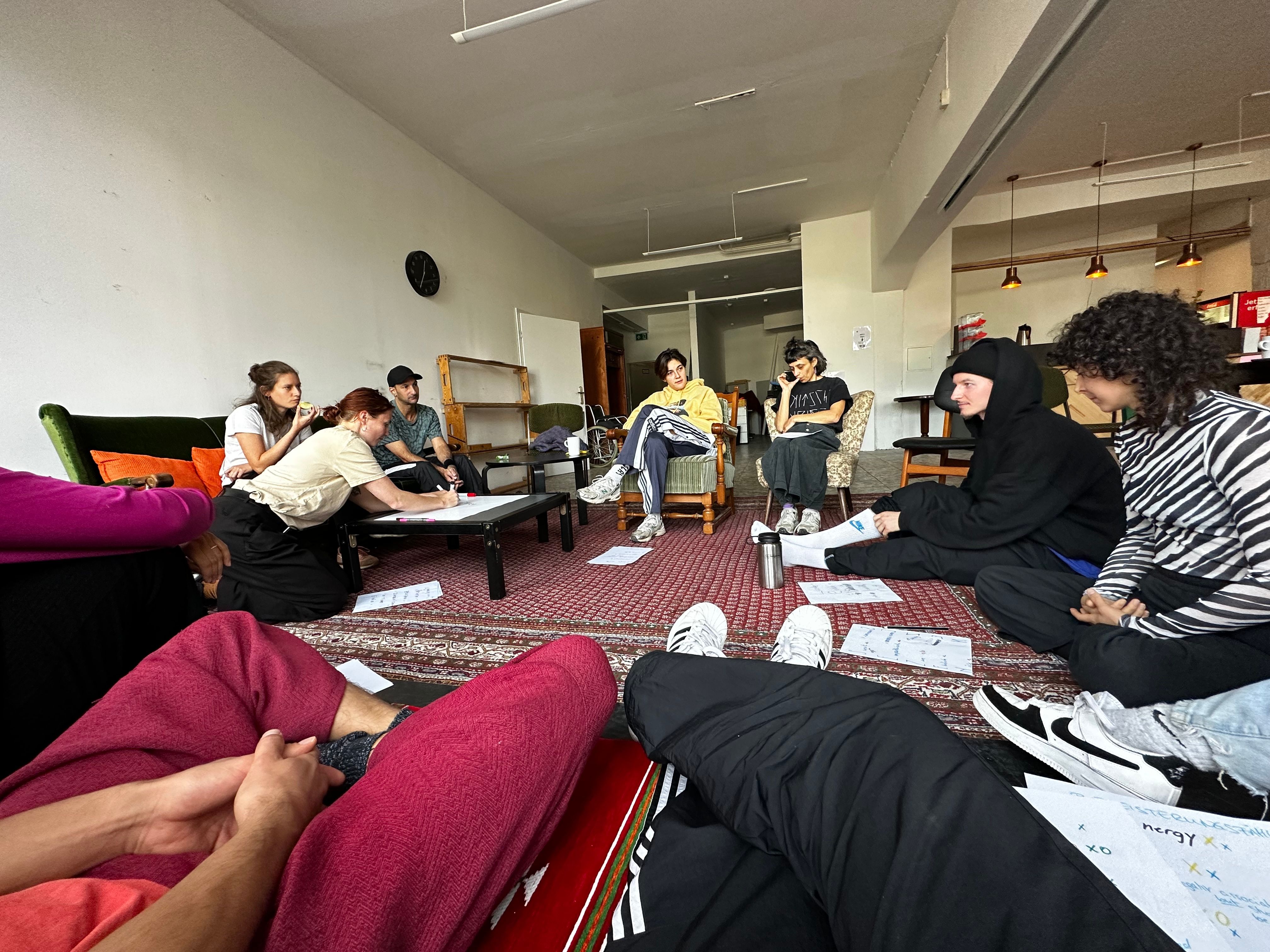 Zu sehen ist eine Gruppe von Menschen in einem Sitzkreis. Es scheint so als würden sie etwas besprechen oder planen.