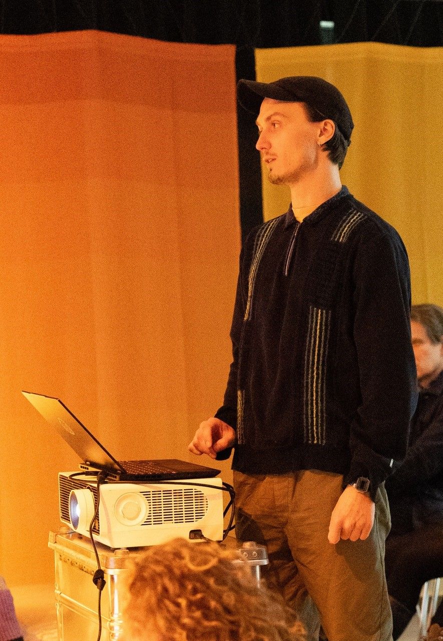 Zu sehen ist Nathan Schönewolf im Ganzkörperprofil. Er trägt einen schwarzen Pullover und eine schwarze Kappe. Vor ihm ist erhöht ein aufgeklappter Laptop zu sehen.