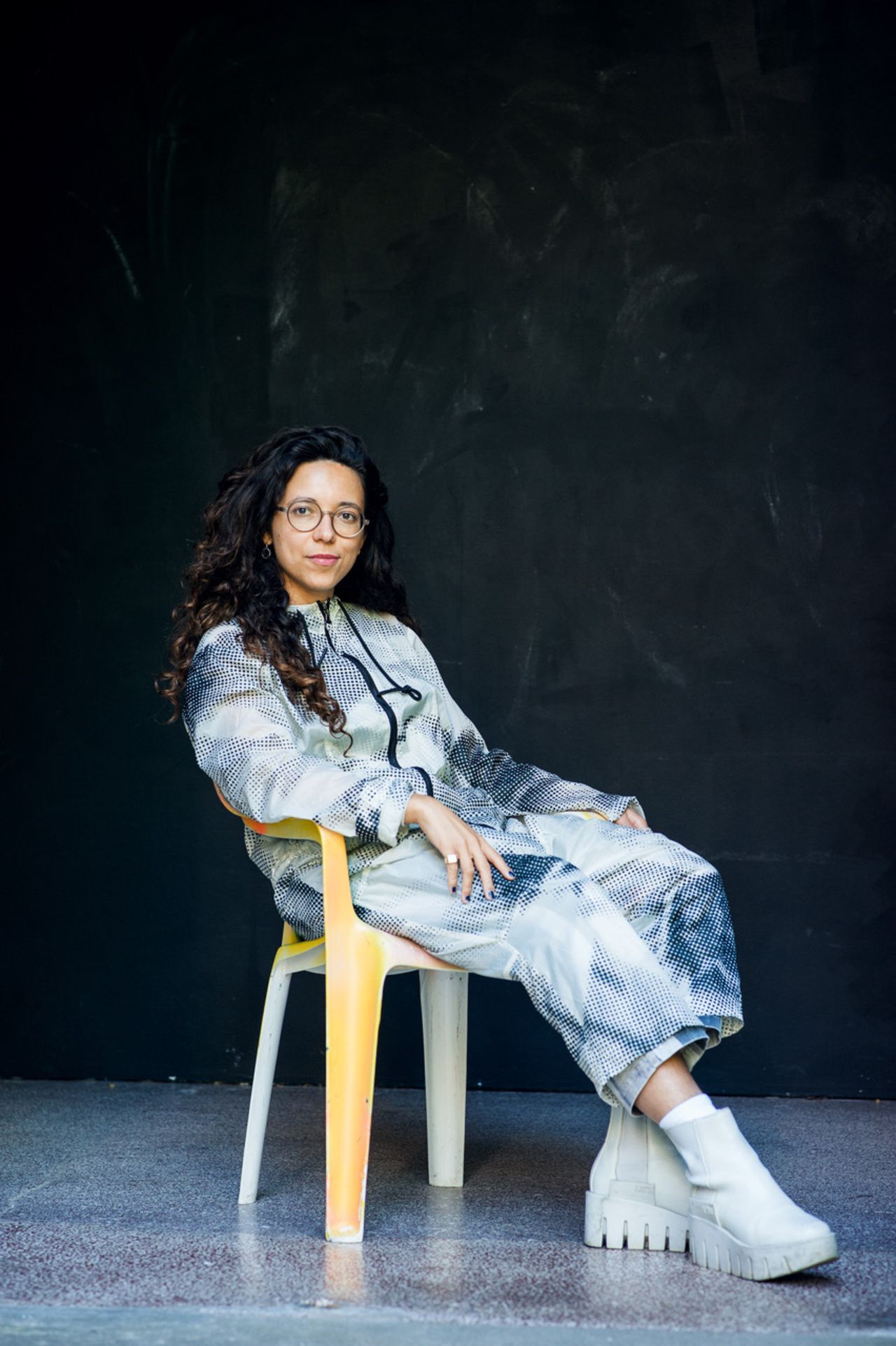 Das Foto zeigt Dr. Lea Luka Sikau im Ganzkörperportrait. Sie trägt einen sehr hellgrünen Ganzkörperanzug mit einem Pixelmuster in schwarz, sitzt auf einem Orange-weißen Plastikstuhl und streckt ein Bein aus. Ihr Blick ist freundlich zur Kamera gewandt mit einem leichten Lächeln. Dr. Sikau trägt eine runde Brille.