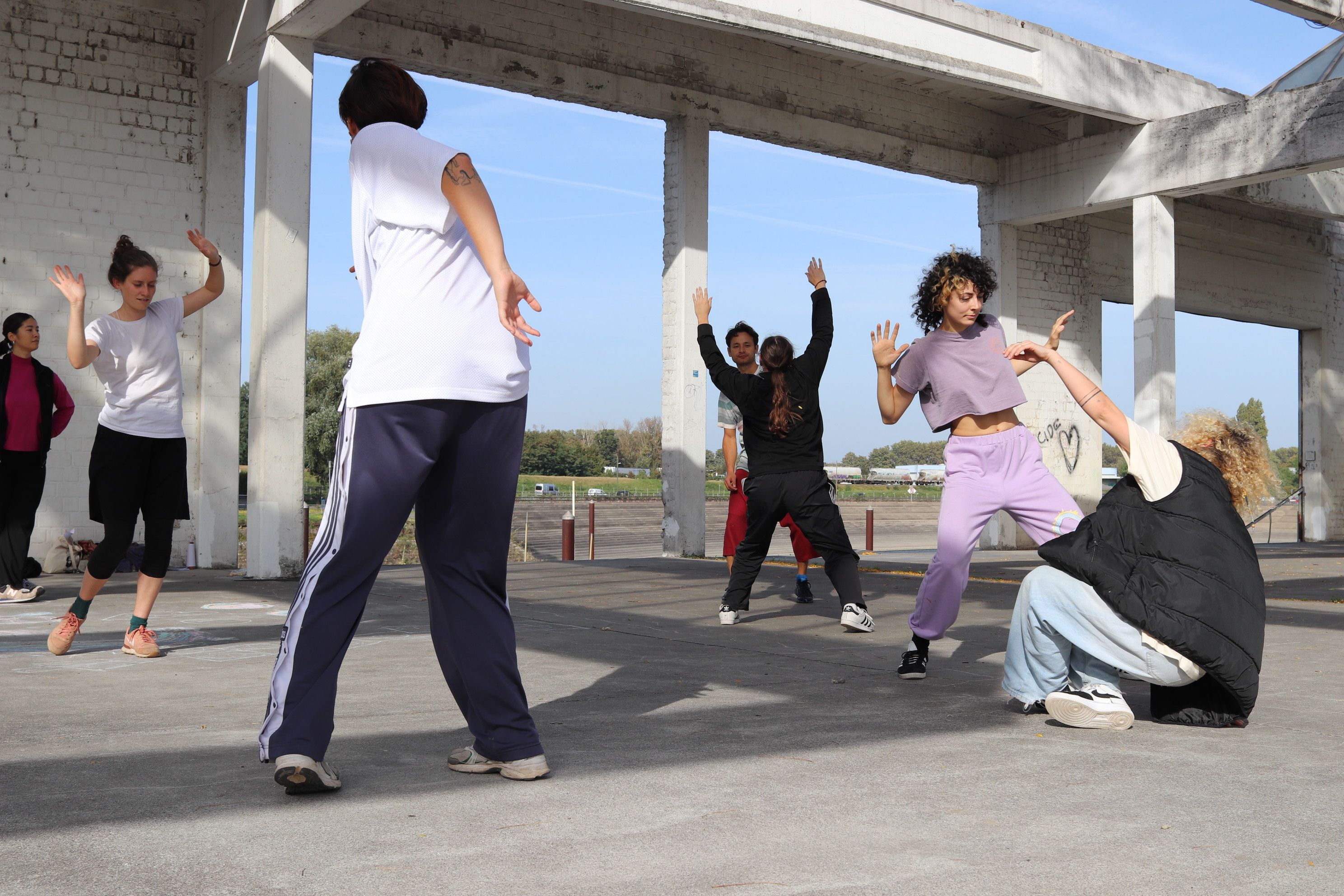 Junge Menschen tanzen auf betoniertem Boden, ein Spiel aus Licht und Schatten, eine Art Gerüst aus Betonträgern im Hintergrund