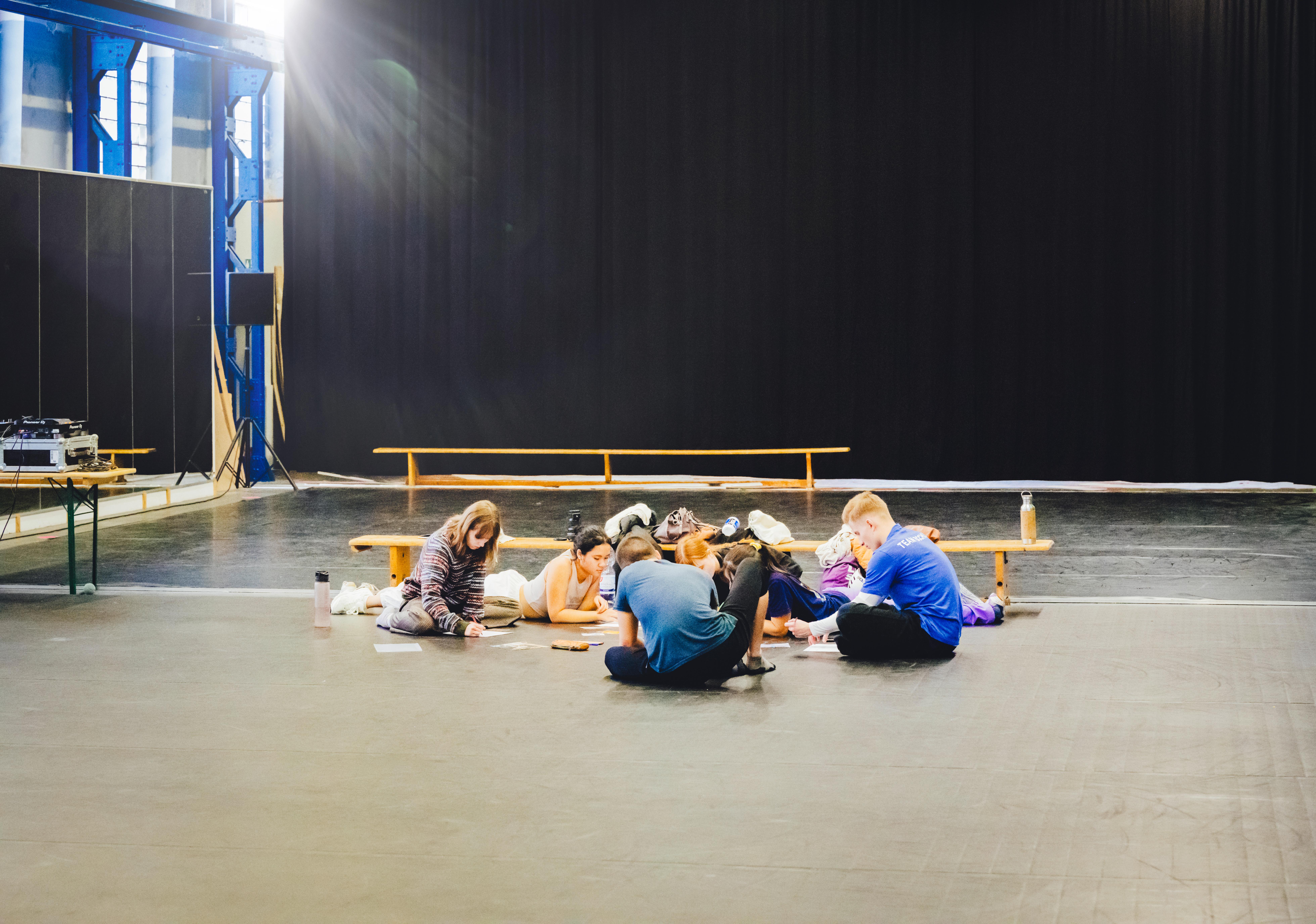 Zu sehen ist eine Gruppe von jungen Menschen die in einem Tanzsaal auf dem Boden zusammensitzt. Es wirkt so als würden sie was schreiben.