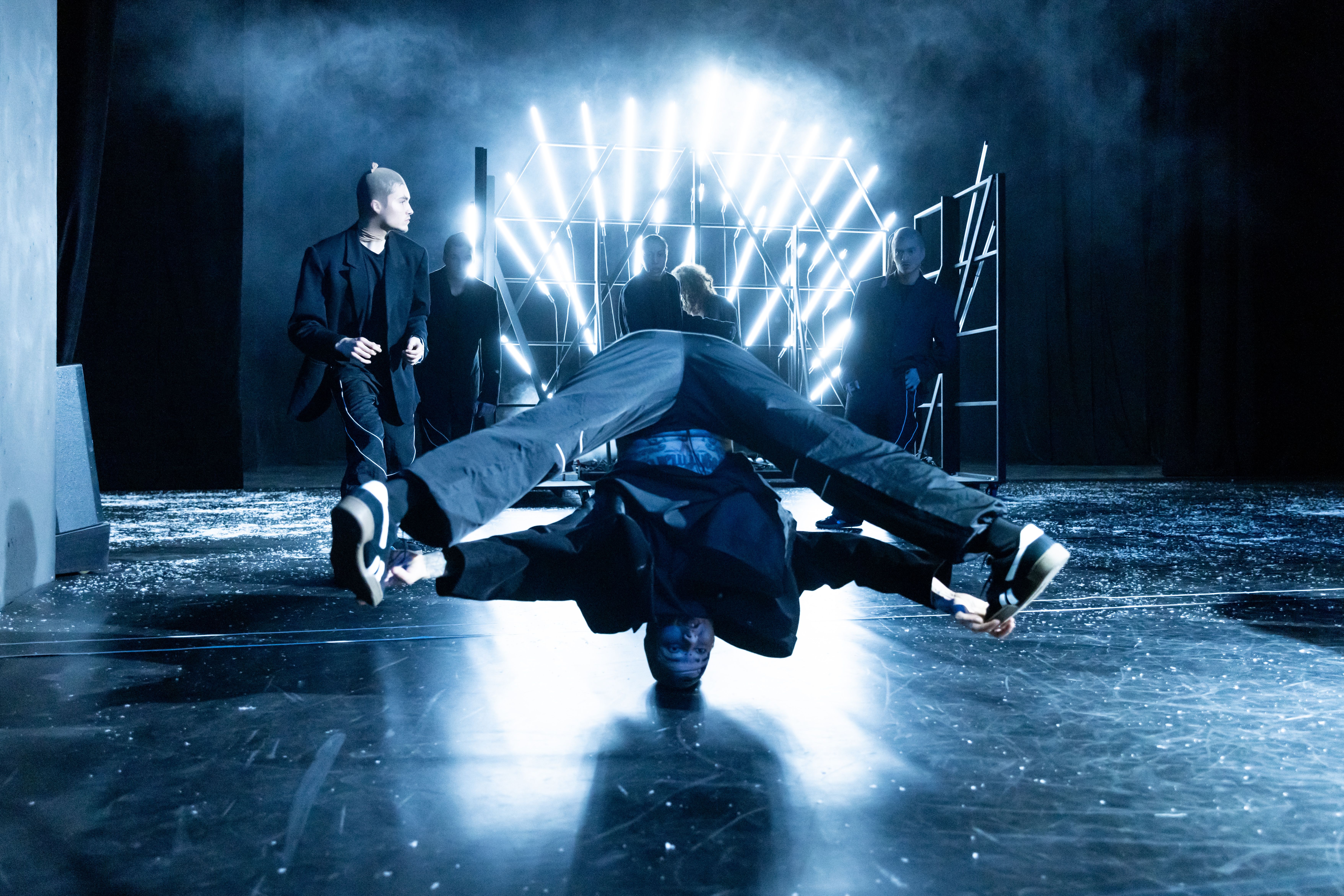 Auf dem Foto sind die Tänzer*innen des Urban Arts Ensembles zu sehen. Zentral ist eine Person zu sehen, die einen Head-Spin macht. Im Hintergrund befinden sich 5 weitere Tänzer*innen.