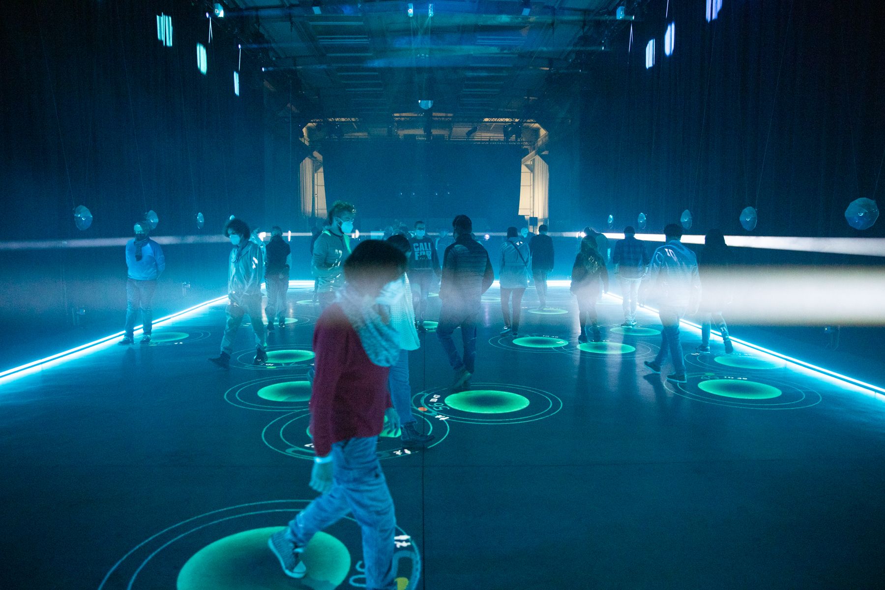 Mehrere Menschen laufen in einem abgedunkelten Raum auf einer Bühnenfläche. Jede Person ist mit einer hellgrün leuchtenden Bodenmarkierung verbunden. Der rechteckige Raum ist am Boden seitlich durch ein LED-Band gesäumt.