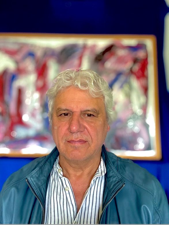 Jurymitglied Christian Strüder in einer blauen geöffneten Jacke und einem gestreiften Hemd vor einem bunten verschwommenen Hintergrund, der ein Bild in einer Ausstellung vermuten lässt.