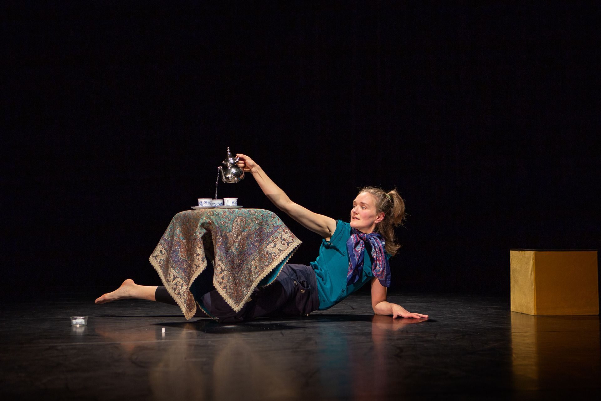 Künstler:in, die auf einer Bühne liegt und Tee in Tassen einschenkt