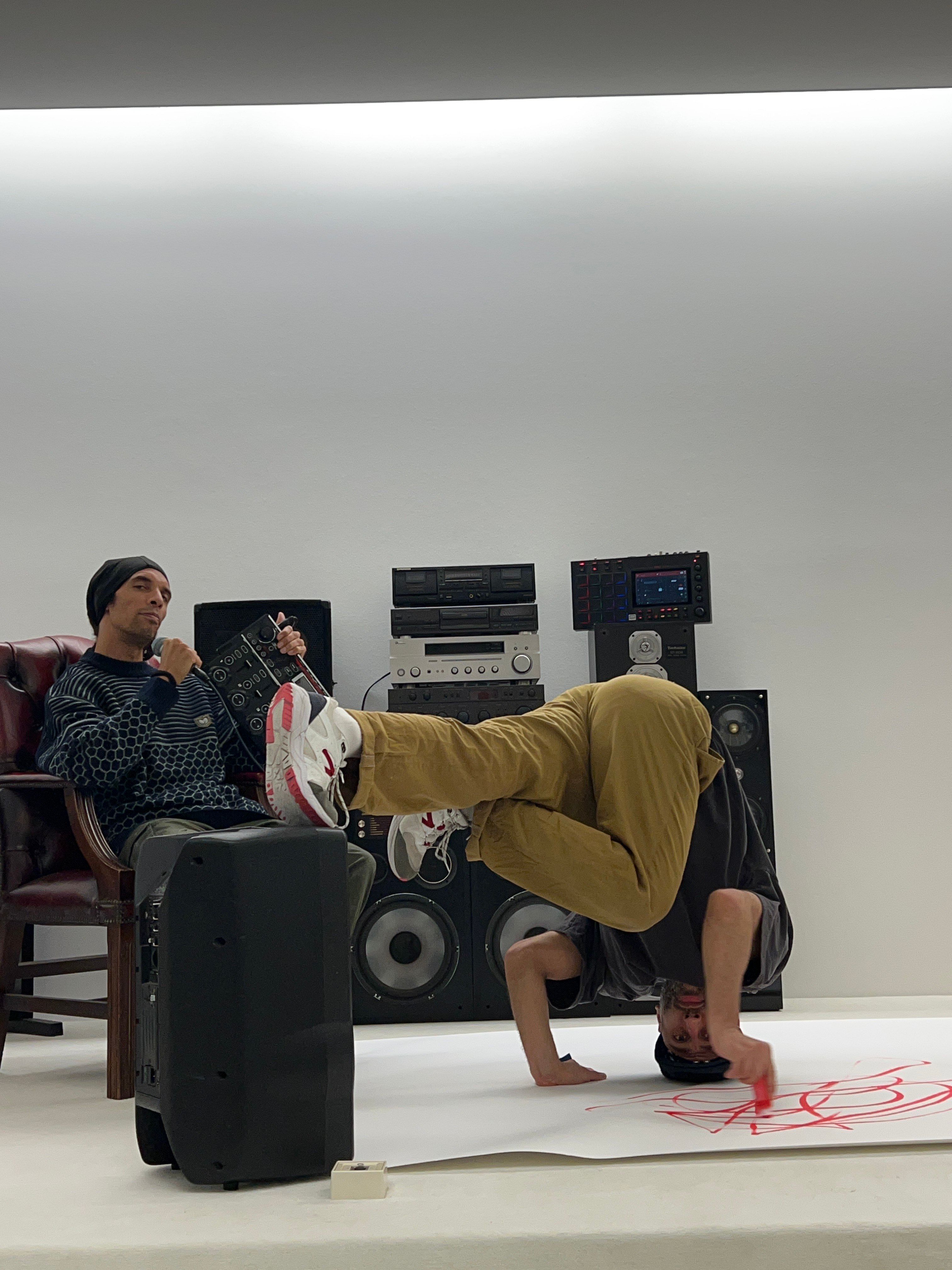 Zu sehen sind zwei Personen. Eine Person sitzt auf einem Sessel und hat ein Mikro sowie ein kleines Mischpult in der Hand. Die andere Person befindet sich auf dem Boden in einer Pose.