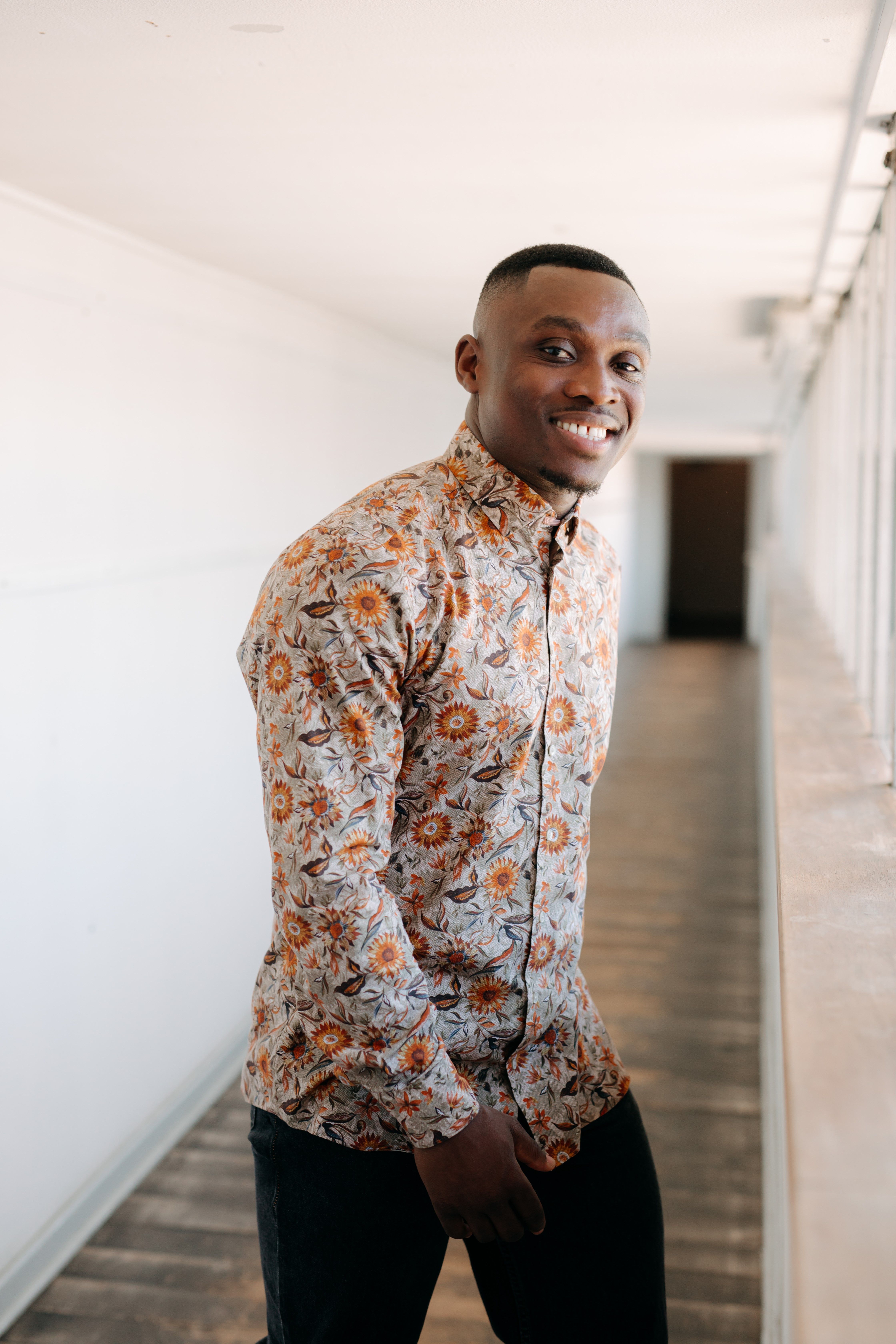 Zu sehen ist der Künstler Kwame Osei, der in einem Blumenhemd in Herbstfarben lächelnd in einem Flur steht.