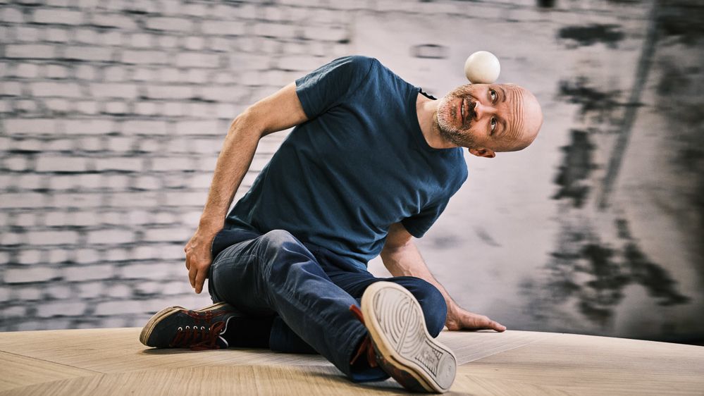 Das Bild zeigt eine Person, die auf einer Holzplatte sitzt und den Kopf schräg hält. Auf dem Ohr der Person, das nach oben zeigt liegt ein faustgroßer Ball.