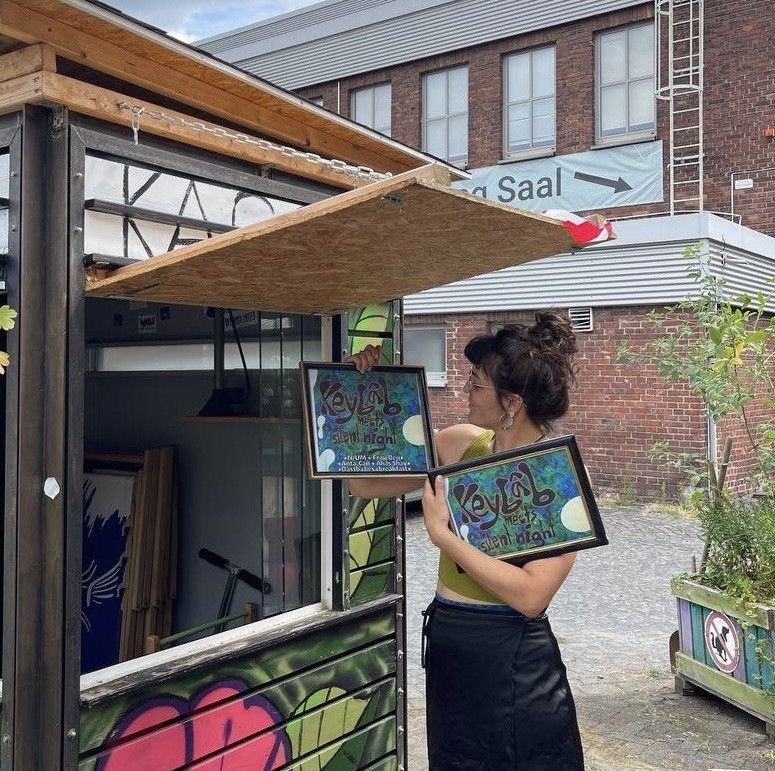 A person holds up two picture frames with photos in front of an open booth. She looks at them. The photo was taken outside.
